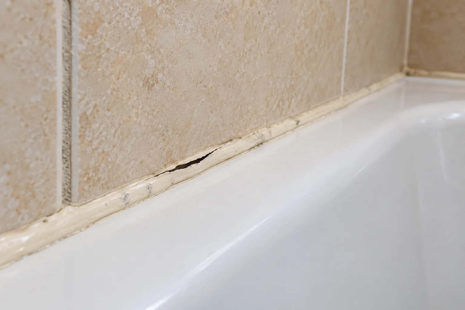 Aged shower sealant with a split and detached edge between beige wall tile and a white bathtub rim