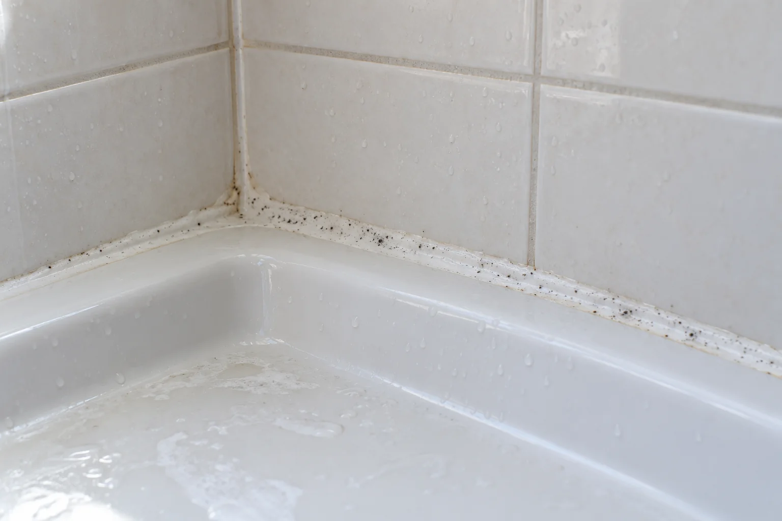 Dark spots and soap residue along white shower caulk where tiled walls meet the shower tray